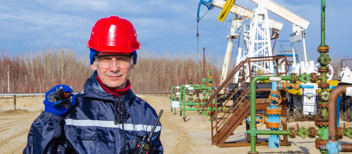 Portrait of man engineer in the oil field wearing red helmet and work clothes holding wrenches in his hand and radio in jacket pocket. Blurry pump jack and wellhead background. Oil and gas concept.